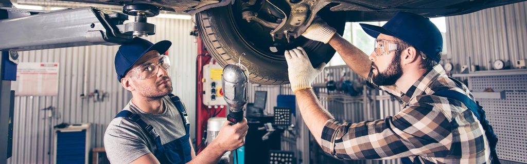 Two mechanics working on a car's suspension system, one using a wrench and the other operating a power tool.